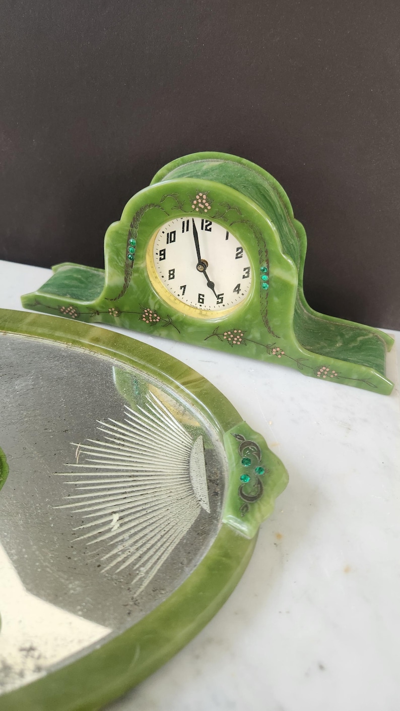 May include: A vintage green clock with a white clock face and black hands. The clock has a decorative green frame with gold and green accents. A matching green tray with a sunburst design is in the foreground.