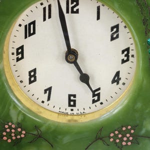 May include: A vintage green clock with a white face and black numerals. The clock has a gold-coloured trim and black hands. Decorative pink floral accents are on the clock face and frame. The words "MADE IN U.S.A." are printed on the clock face.