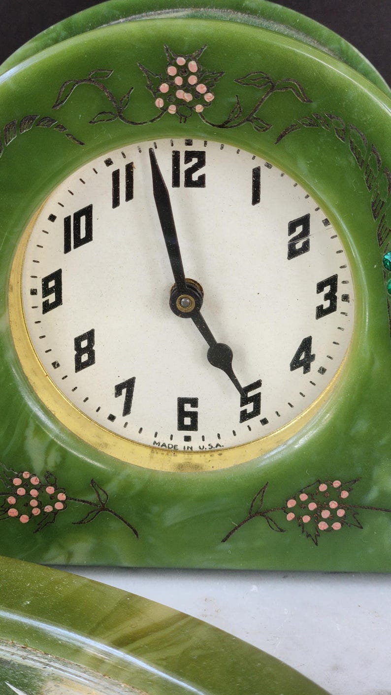 May include: A vintage green clock with a white face and black numerals. The clock has a gold-coloured trim and black hands. Decorative pink floral accents are on the clock face and frame. The words "MADE IN U.S.A." are printed on the clock face.