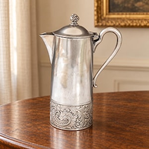 May include: A tall silver-toned metal coffee pot featuring a smooth, polished upper body and a base decorated with intricate, embossed floral and scrollwork patterns. It includes a curved handle, a pointed spout, and a lid with a small finial.