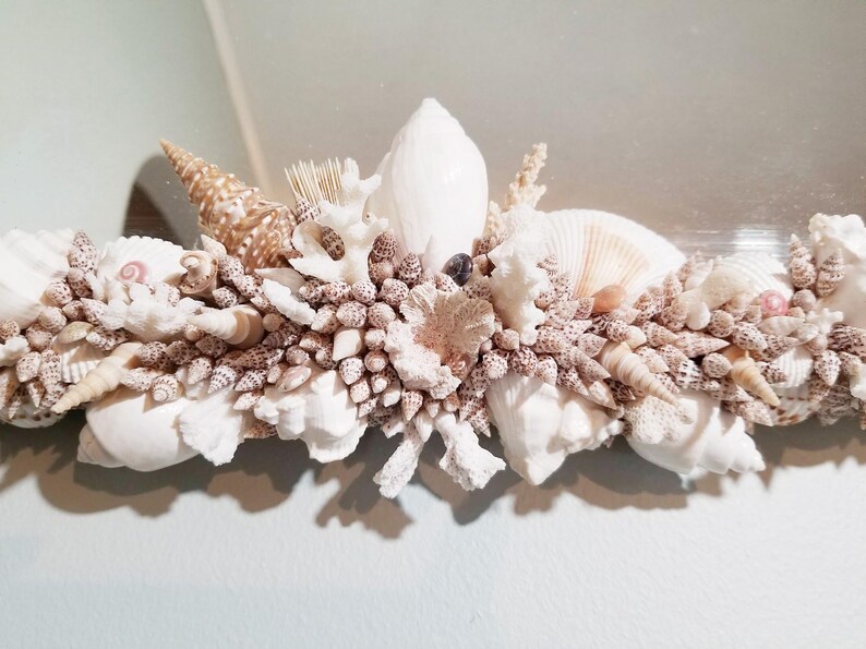 May include: A decorative mirror frame adorned with a variety of seashells, coral, and small conch shells. The shells are primarily white, beige, and brown, creating a coastal theme. The frame is ideal for a bathroom or beach-themed decor.