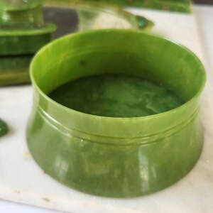 May include: A green Art Deco vanity set, comprising a round container, a small lid, and a clock. The set is made of a translucent, jade-coloured material. The clock has a white face with black numerals and hands.