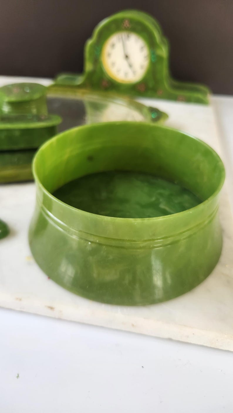 May include: A green Art Deco vanity set, comprising a round container, a small lid, and a clock. The set is made of a translucent, jade-coloured material. The clock has a white face with black numerals and hands.