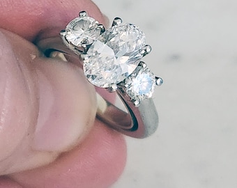 May include: A silver ring featuring an oval-cut center stone flanked by two smaller round stones and two additional round stones. The ring is held up against a light background, showcasing the sparkle of the gemstones.