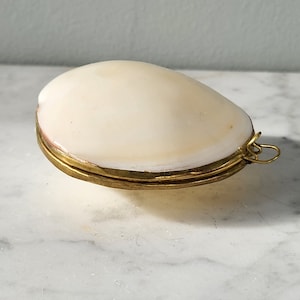 May include: A small, white seashell with a gold metal rim and a small loop on the side. The seashell is closed and sitting on a white marble surface.