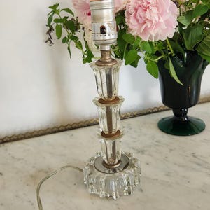 May include: A clear glass table lamp base featuring a tiered, fluted design with a metallic socket at the top. The lamp has a circular, scalloped glass base and includes a thin, transparent electrical cord extending from the bottom.