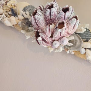 May include: A decorative mirror frame adorned with various seashells, coral, and a large, textured, barnacle-like centerpiece in shades of white and burgundy. The frame is embellished with small, light-colored seashells and coral.