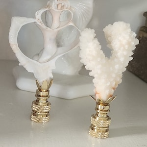 May include: Two gold lamp finials with a seashell and coral design. The seashell is white with a pink hue and the coral is white. The finials are on a white surface.