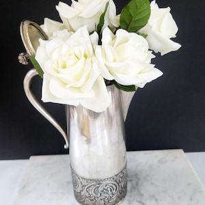 Op de afbeelding: Een zilveren kan met een handvat en een decoratieve basis gevuld met witte rozen en groene bladeren. De kan staat op een wit marmeren oppervlak tegen een zwarte achtergrond. De rozen staan in volle bloei.