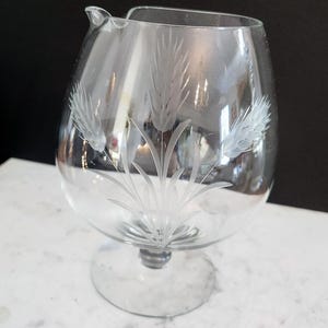 May include: Clear glass creamer with an etched wheat stalk design. The creamer has a rounded body, a small spout, and a pedestal base. The etched design is delicate, adding elegance. The creamer is suitable for serving milk or cream.