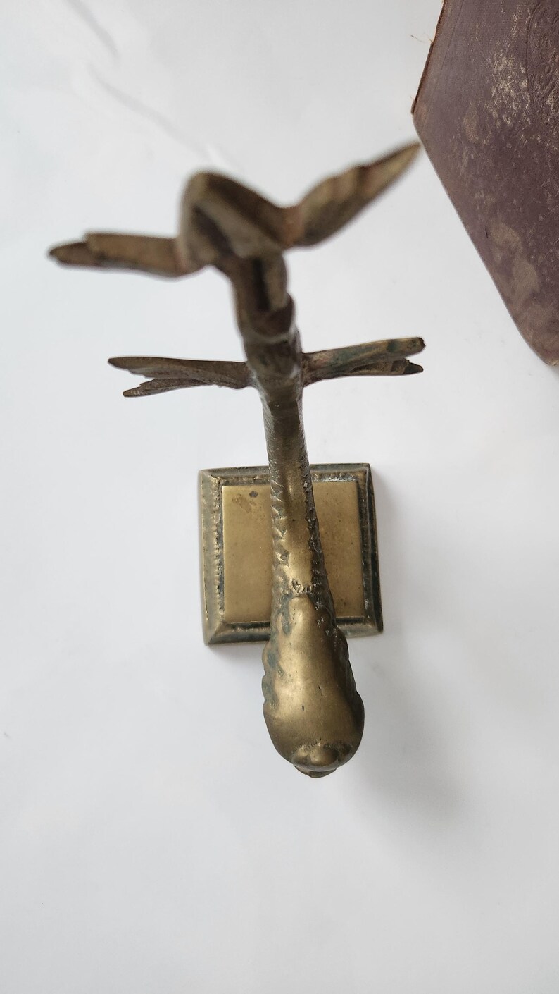 May include: A vintage brass sculpture of a bird in flight. The sculpture has a rectangular base and a detailed bird form with outstretched wings. The brass has a warm, aged patina, suggesting its antique nature. The bird's form is detailed and elegant.