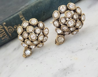 May include: A pair of vintage-style earrings. Each earring features a cluster of small, round, clear crystals set in a gold-toned metal. The crystals are arranged in a floral or circular pattern, creating a sparkling effect. The earrings have a post-back closure.