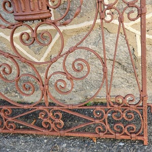 Pu&ograve; includere: Primo piano di una sezione di un cancello in ferro battuto arrugginito e ornato. Il cancello presenta un'intricata lavorazione a volute ed elementi decorativi, tra cui spirali e motivi floreali. Il colore marrone-rossastro del cancello suggerisce l'et&agrave; e l'esposizione agli agenti atmosferici.