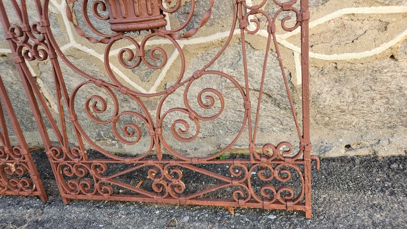 Pu&ograve; includere: Primo piano di una sezione di un cancello in ferro battuto arrugginito e ornato. Il cancello presenta un'intricata lavorazione a volute ed elementi decorativi, tra cui spirali e motivi floreali. Il colore marrone-rossastro del cancello suggerisce l'et&agrave; e l'esposizione agli agenti atmosferici.