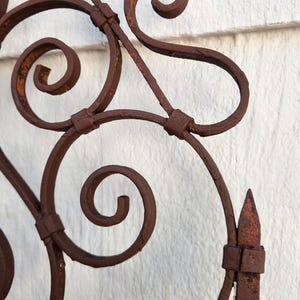 Pu&ograve; includere: Primo piano di una sezione di un cancello decorativo in ferro battuto. Il cancello presenta un'intricata voluta e un pinnacolo appuntito. Il ferro ha una finitura marrone arrugginita, che suggerisce l'et&agrave; e l'uso all'aperto. Lo sfondo &egrave; un muro bianco strutturato.