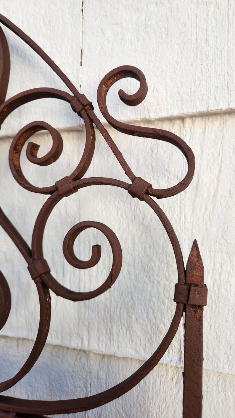 Pu&ograve; includere: Primo piano di una sezione di un cancello decorativo in ferro battuto. Il cancello presenta un'intricata voluta e un pinnacolo appuntito. Il ferro ha una finitura marrone arrugginita, che suggerisce l'et&agrave; e l'uso all'aperto. Lo sfondo &egrave; un muro bianco strutturato.