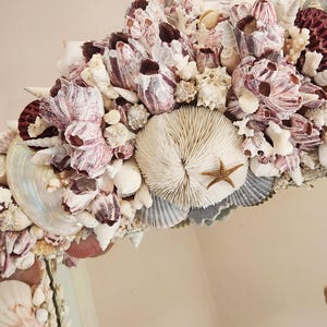May include: A decorative mirror frame adorned with a variety of seashells, barnacles, and coral. The frame features a mix of colors, including white, pink, and brown, with a small starfish accent. The design creates a coastal or nautical theme.