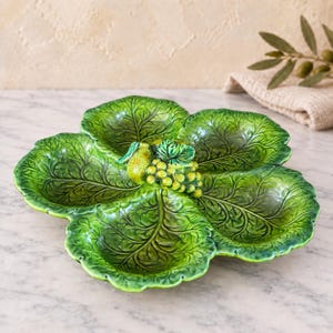 May include: A ceramic serving dish shaped like four overlapping green cabbage leaves. The center features a decorative cluster of sculpted green grapes and a single pear. The dish has a glossy finish with detailed leaf veins and scalloped edges.
