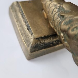 May include: Close-up of a bronze-colored decorative object. The object has a rectangular base and a curved, textured upper portion. The surface shows signs of age and wear, with darker areas along the edges.