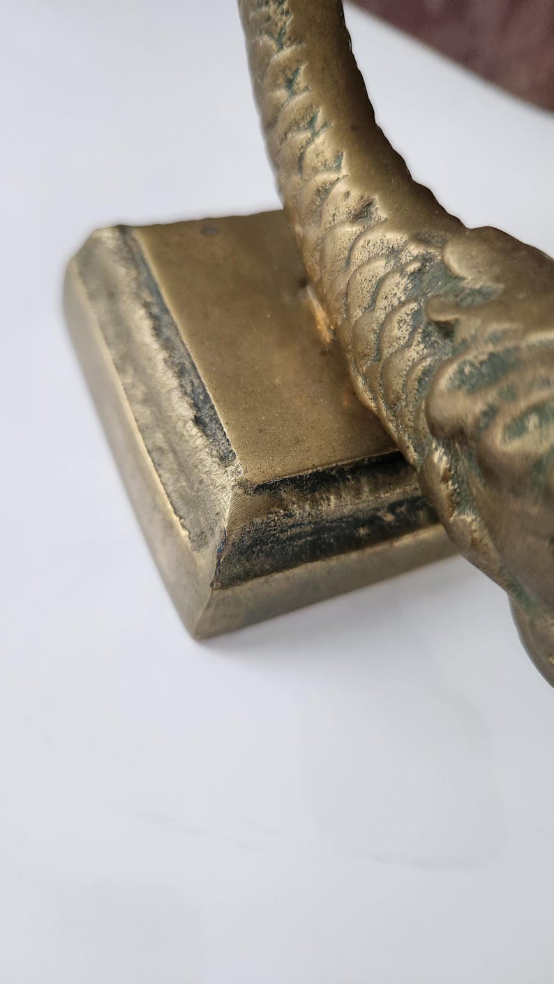 May include: Close-up of a bronze-colored decorative object. The object has a rectangular base and a curved, textured upper portion. The surface shows signs of age and wear, with darker areas along the edges.