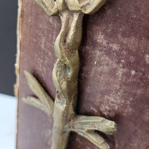 May include: A vintage brass bookend with a stylized, abstract design. The bookend features a twisted central element and outstretched, wing-like details. The brass has an aged patina, and the bookend is set against a burgundy book.