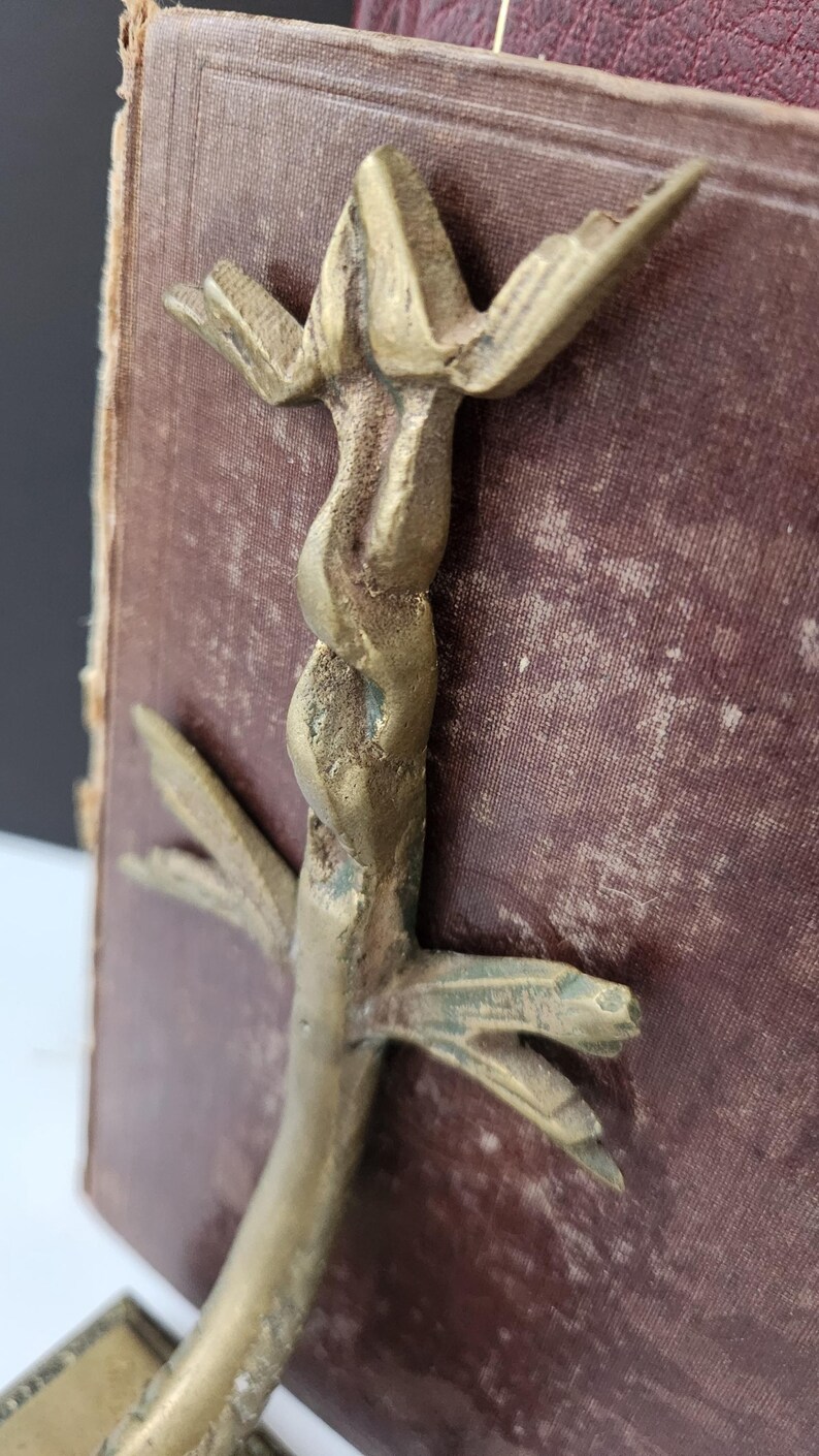 May include: A vintage brass bookend with a stylized, abstract design. The bookend features a twisted central element and outstretched, wing-like details. The brass has an aged patina, and the bookend is set against a burgundy book.
