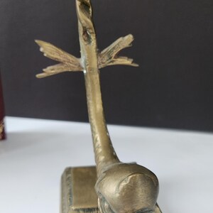 May include: A vintage brass sculpture featuring a stylized dolphin design. The sculpture has a square base and a twisted stem that supports a fin-like element. The dolphin's head is detailed with a textured surface.
