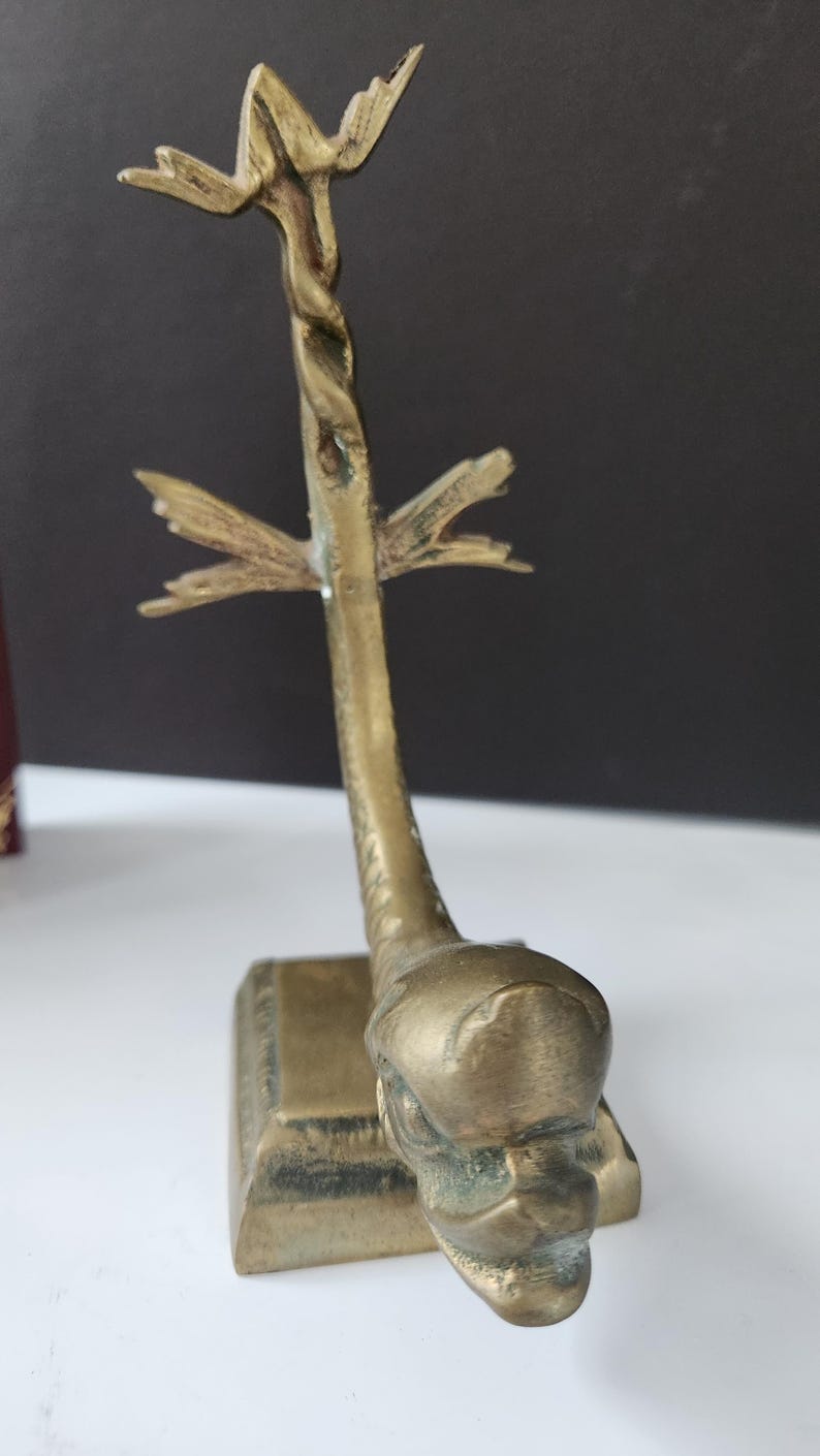 May include: A vintage brass sculpture featuring a stylized dolphin design. The sculpture has a square base and a twisted stem that supports a fin-like element. The dolphin's head is detailed with a textured surface.