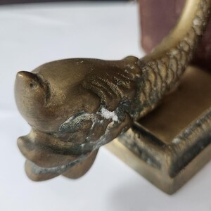 May include: A vintage brass bookend featuring a detailed fish design. The bookend has a rectangular base and a curved, textured body. The fish head is prominent, with visible scales and fins. The brass has an aged patina.