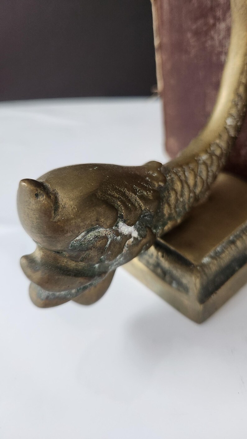 May include: A vintage brass bookend featuring a detailed fish design. The bookend has a rectangular base and a curved, textured body. The fish head is prominent, with visible scales and fins. The brass has an aged patina.
