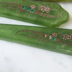May include: Close-up of green vintage serving utensils. Each piece features an etched design with a floral motif, brown accents, and three green gems. The utensils are displayed on a white surface.