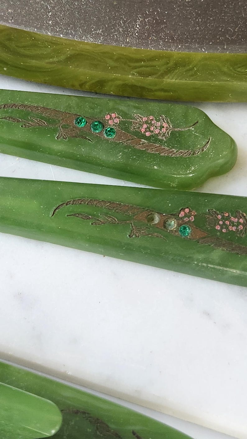 May include: Close-up of green vintage serving utensils. Each piece features an etched design with a floral motif, brown accents, and three green gems. The utensils are displayed on a white surface.