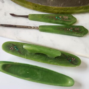May include: A set of vintage green manicure tools, including cuticle pushers and a nail file, with floral detailing and rhinestones. The tools are displayed on a white surface, indicating a focus on beauty and grooming.
