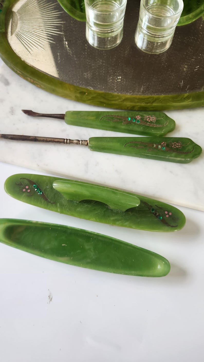 May include: A set of vintage green manicure tools, including cuticle pushers and a nail file, with floral detailing and rhinestones. The tools are displayed on a white surface, indicating a focus on beauty and grooming.