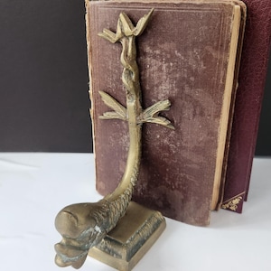 May include: A vintage brass bookend featuring a stylized fish design with a central stem and leaf-like accents. The bookend is positioned in front of two antique books with burgundy covers, creating a classic aesthetic.