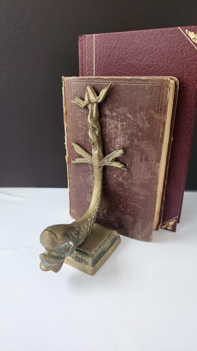 May include: A vintage brass bookend featuring a stylized fish design with a central stem and leaf-like accents. The bookend is positioned in front of two antique books with burgundy covers, creating a classic aesthetic.