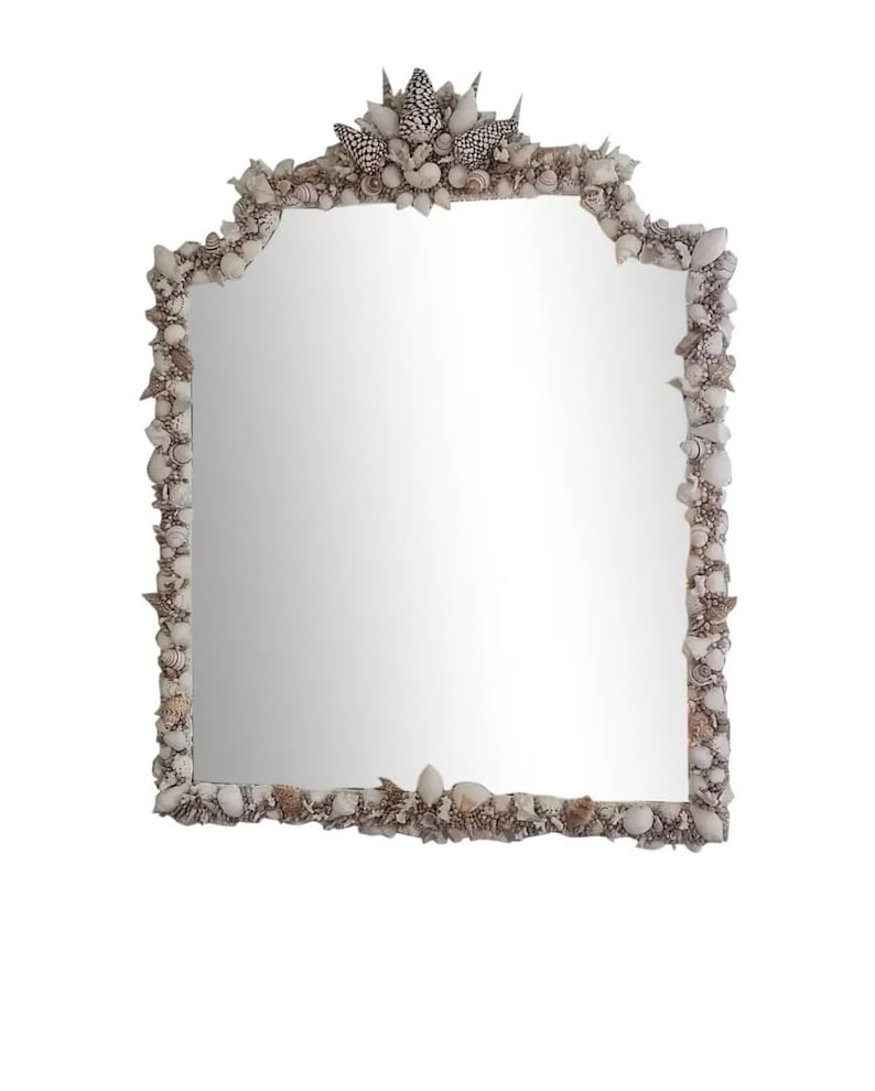 May include: A rectangular mirror with a decorative frame made of various seashells. The frame is a mix of white, beige, and black shells, creating a textured border around the reflective glass. The mirror has a unique, arched top.