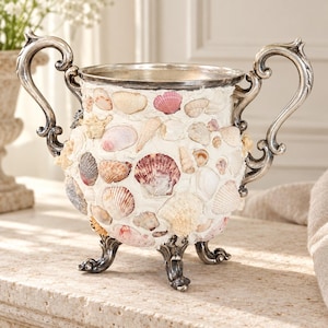 May include: A decorative urn featuring a white textured surface embedded with various seashells in shades of pink, tan, and white. The vessel has two ornate silver-toned metal handles and stands on three matching silver-toned metal feet.