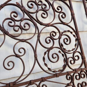 Pu&ograve; includere: Sezione di cancello antico in ferro battuto con finitura color ruggine. Il design presenta motivi a volute, spirali e un accento floreale in alto. Il cancello &egrave; posto su uno sfondo bianco.