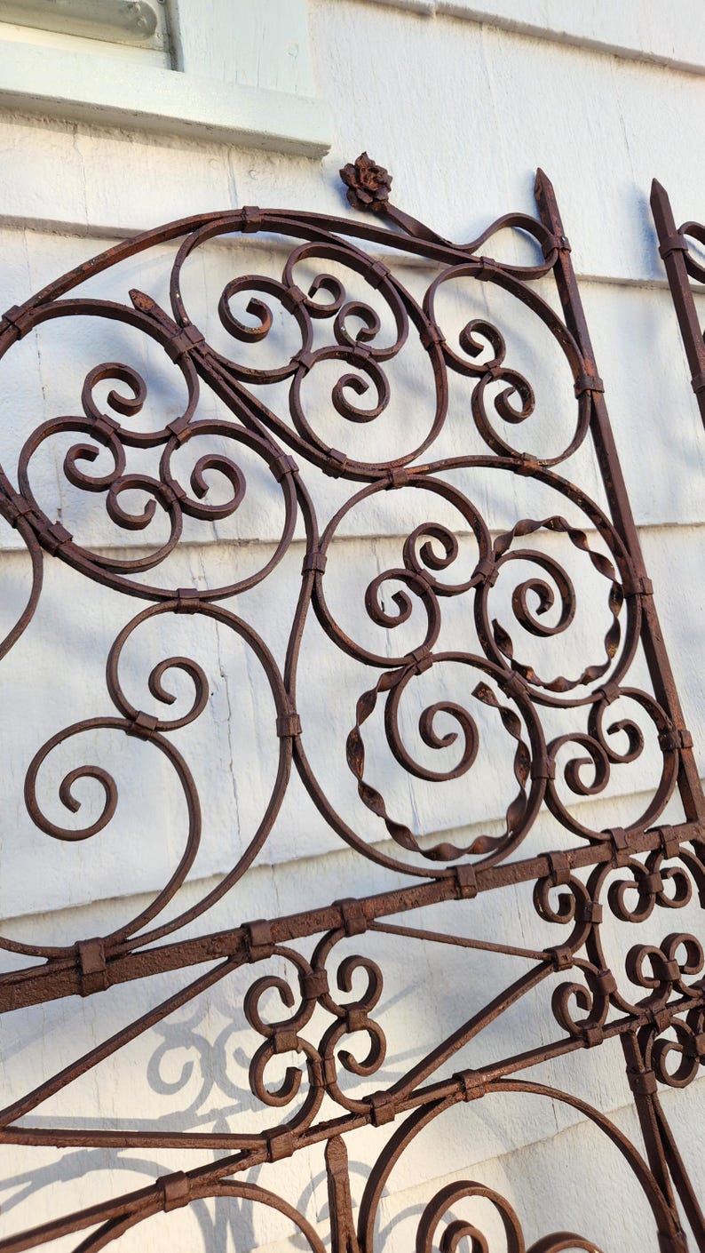 Pu&ograve; includere: Sezione di cancello antico in ferro battuto con finitura color ruggine. Il design presenta motivi a volute, spirali e un accento floreale in alto. Il cancello &egrave; posto su uno sfondo bianco.