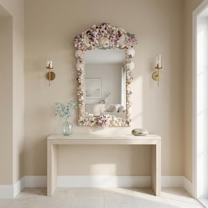 May include: A decorative mirror with a seashell frame hangs above a light-colored wooden console table. The mirror's frame is adorned with various seashells and coral in shades of white, pink, and beige. Two gold sconces flank the mirror, and a glass vase with greenery sits on the table.