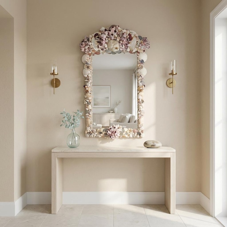May include: A decorative mirror with a seashell frame hangs above a light-colored wooden console table. The mirror's frame is adorned with various seashells and coral in shades of white, pink, and beige. Two gold sconces flank the mirror, and a glass vase with greenery sits on the table.