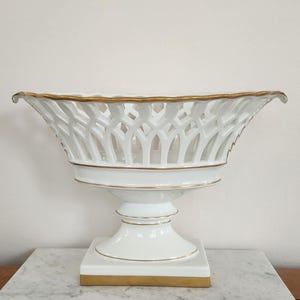 May include: White porcelain fruit bowl with a lattice design and gold trim. The bowl sits on a pedestal base with a square foot, also trimmed in gold. A decorative piece for the home.