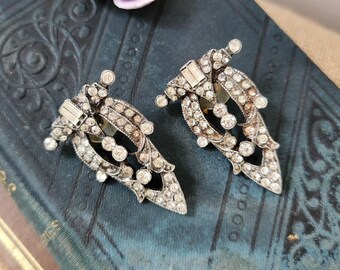 May include: A pair of vintage silver-toned clip-on earrings, encrusted with clear rhinestones. The earrings have a leaf-like design with a central open space. A small, light purple flower is also visible.