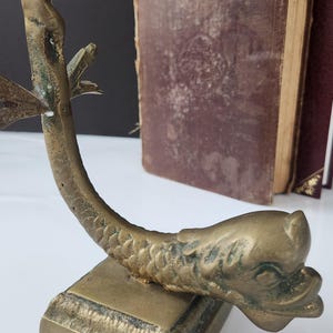 May include: A vintage brass bookend in the shape of a fish, with a detailed scale pattern and a square base. The bookend is positioned in front of two antique books with worn, burgundy covers.