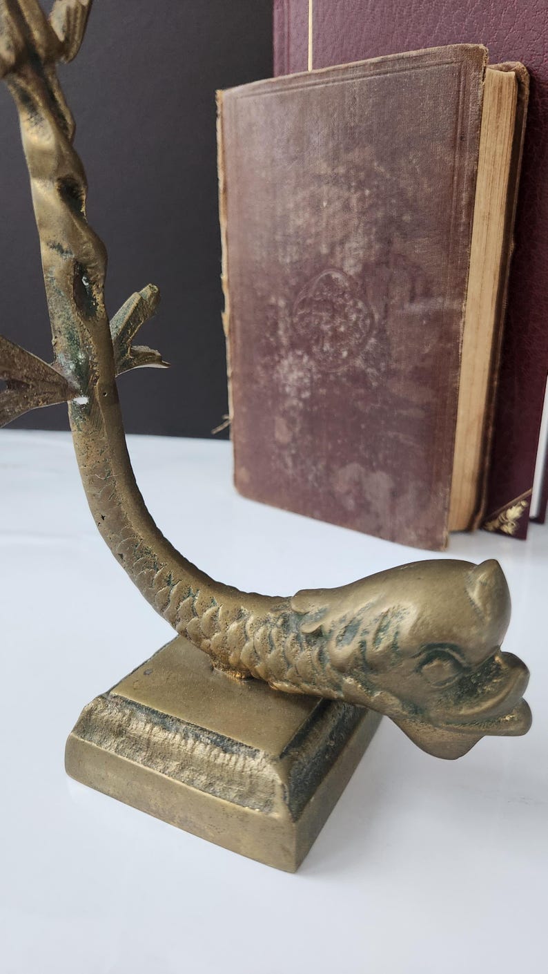 May include: A vintage brass bookend in the shape of a fish, with a detailed scale pattern and a square base. The bookend is positioned in front of two antique books with worn, burgundy covers.