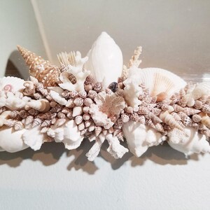 May include: A decorative mirror frame adorned with a variety of seashells, coral, and small conch shells. The shells are primarily white, beige, and brown, creating a coastal theme. The frame is ideal for a bathroom or beach-themed decor.