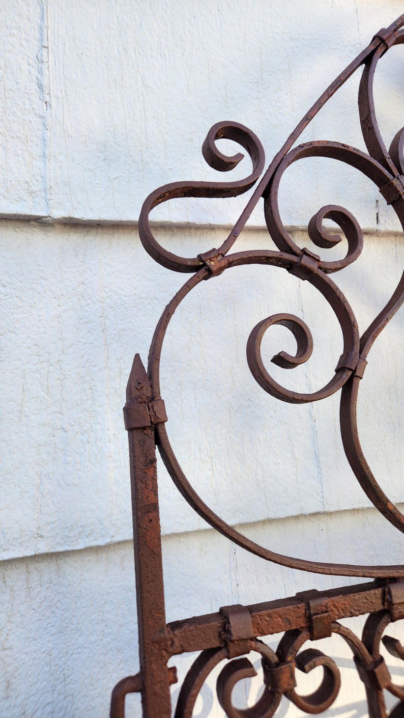 Pu&ograve; includere: Primo piano di una sezione di un cancello decorativo in ferro battuto. Il cancello presenta volute ornate e una finitura marrone arrugginita. Il design include linee curve e accenti appuntiti su sfondo bianco.