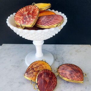 May include: A collection of seashell-shaped trinket boxes with gold-colored trim. The boxes are in shades of red, orange, and yellow, and are displayed in a white, footed bowl and on a marble surface.