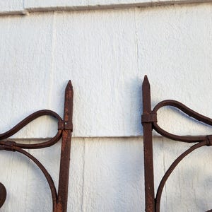 Pu&ograve; includere: Primo piano di una sezione di un cancello in ferro battuto con finitura marrone ruggine. Il cancello presenta volute decorative, pinnacoli appuntiti e accenti floreali. Lo sfondo &egrave; una parete di legno bianco.